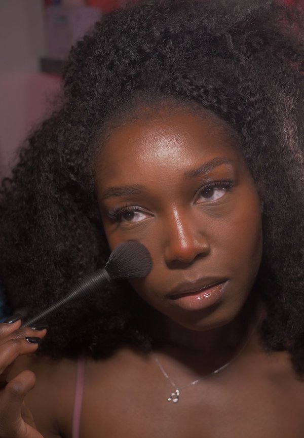 Giovane donna con capelli ricci che applica il trucco con un pennello sulla guancia, concentrata sul suo riflesso o specchio.