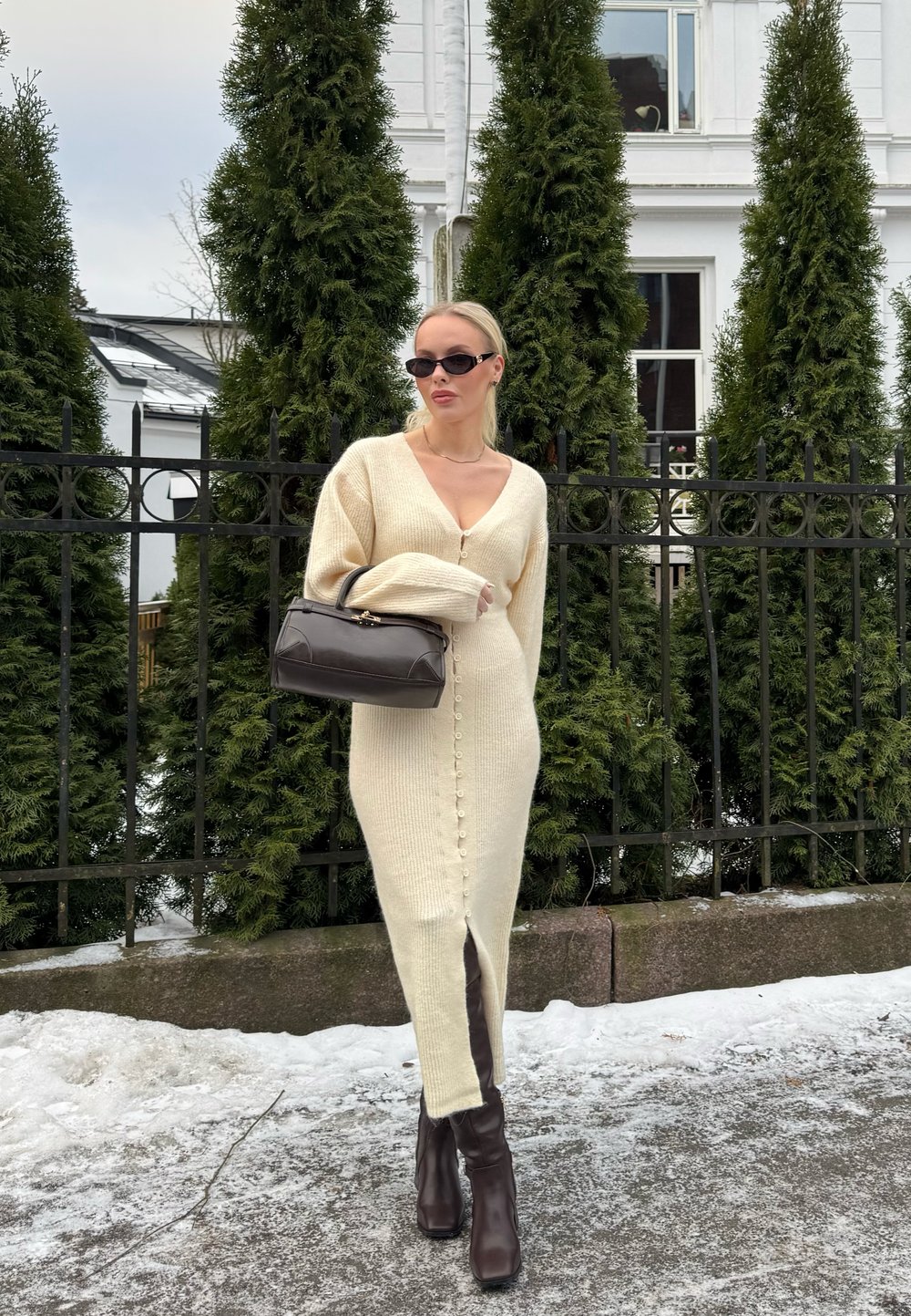 Femme portant une robe en maille crème boutonnée, des bottes foncées, des lunettes de soleil et tenant un sac à main foncé, debout sur un sol enneigé près d'une clôture en métal.