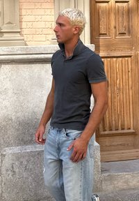 Tmavošedé polo tričko s vyvýšeným golierom skombinované so svetlo modrými, poškodenými džínami. Krátke, textúrované blond vlasy. Pozadie obsahuje drevené dvere a kamennú stenu.