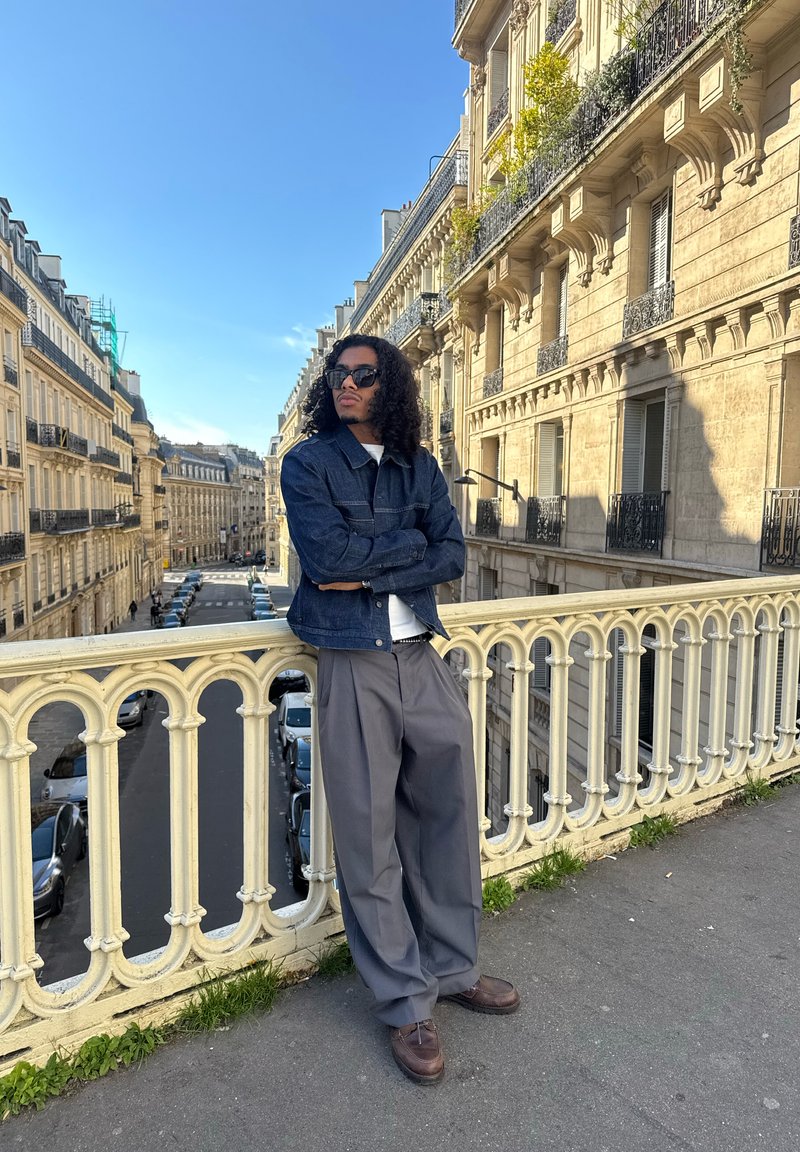 Personne aux cheveux bouclés et lunettes de soleil s'appuie sur une rambarde ornée surplombant une rue parisienne avec des bâtiments classiques en pierre sous un ciel dégagé.