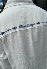 Vit linneskjorta med marinblå broderi av "Finer Things" och dekorativa pilmotiver på baksidan, som visar en avslappnad textur.