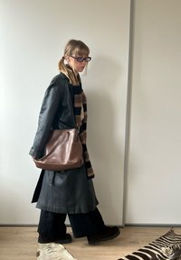 Schwarzer Ledermantel, gestreifte braun-schwarze Schal, braune Lederhandtasche, weite schwarze Hose, dunkle Stiefel, Holzboden, neutraler Hintergrund.
