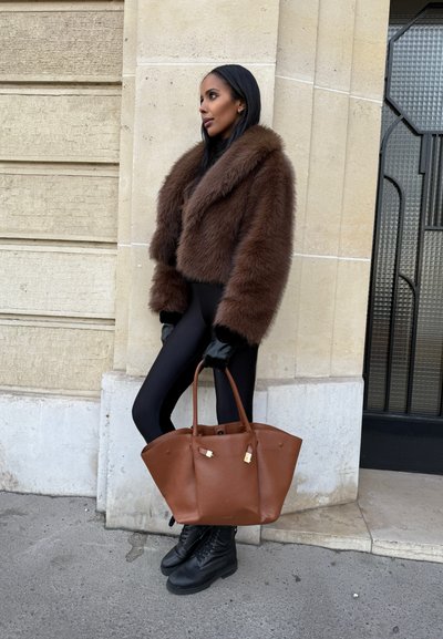 Chaqueta corta de piel sintética marrón, leggings negros, guantes negros, bolso de mano de piel marrón con detalles dorados y botas negras con cordones.