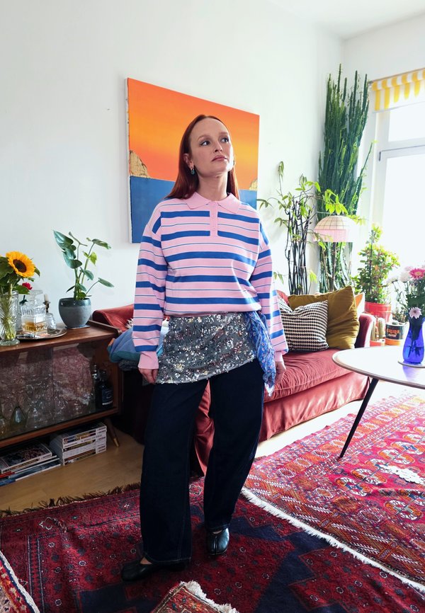 Mujer con el pelo largo y rojo que lleva una camisa de rayas rosa y azul, pantalones negros y una falda con lentejuelas, de pie en una sala de estar colorida con plantas y arte.
