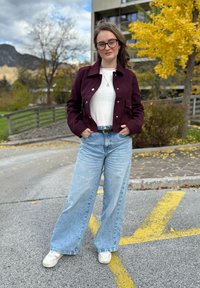 Bordó dzseki, fehér felső, magas derekú, világoskék, bőszárú farmernadrág és fehér sportcipő. Természetes környezet, őszi lombsorral a háttérben.