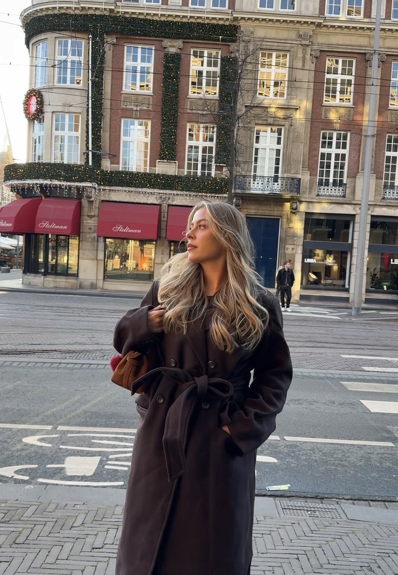 Vrouw met lang blond haar, gekleed in een donkere jas met ceintuur, staat op een straathoek in de stad voor een versierd bakstenen gebouw met rode luifels boven de winkelramen.