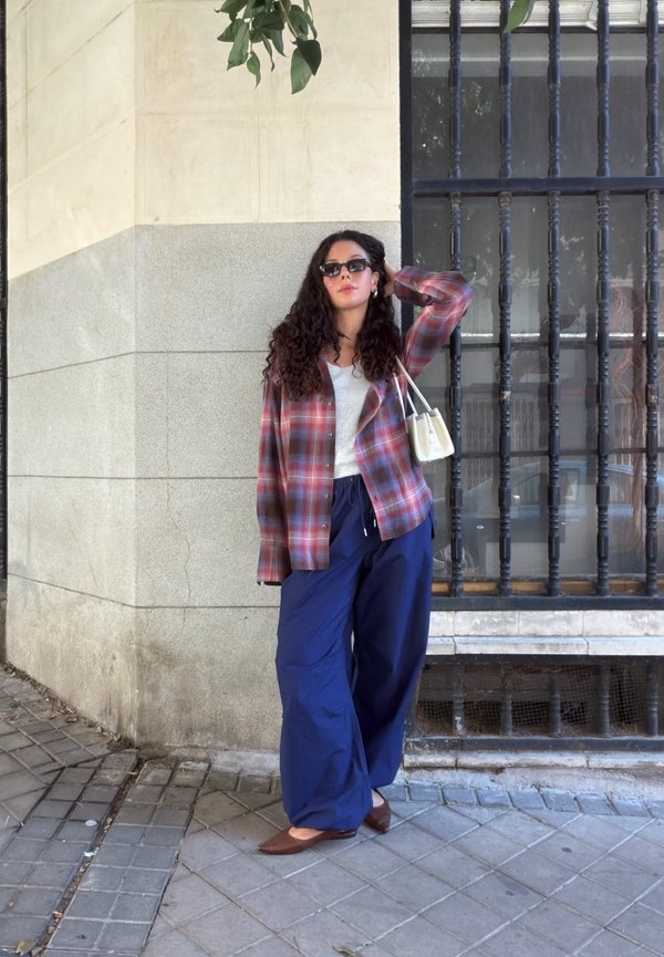Una mujer con gafas de sol y una chaqueta de cuadros posa junto a una ventana enrejada y lleva un bolso blanco al hombro.