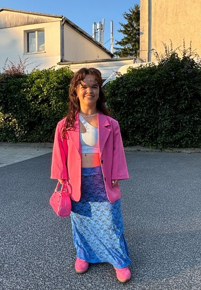 Blazer rosa sobre un top corto blanco, falda azul estampada, zapatos rosas y bolso a juego. Colores vibrantes y texturas en capas. Entorno al aire libre.