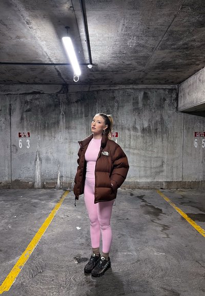 Joven mujer con ropa deportiva rosa y chaqueta acolchada marrón, de pie en un garaje de concreto vacío bajo una luz de techo brillante.