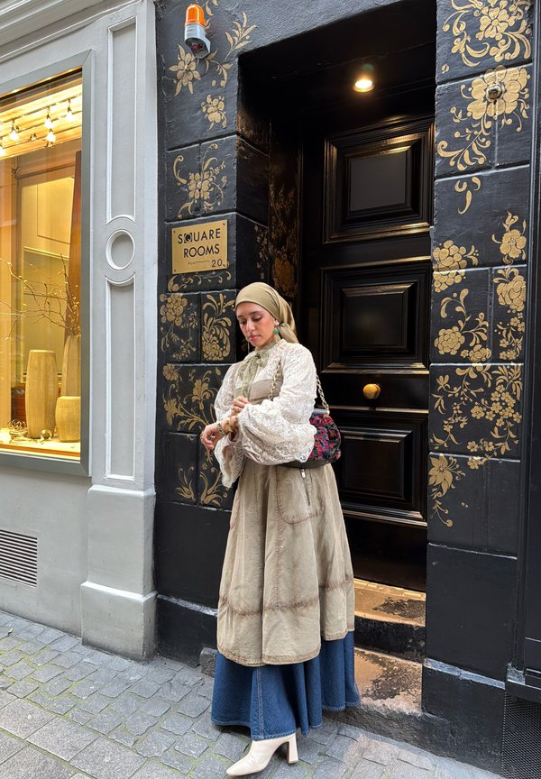 Frau in gestuftem Rock und beigem Kopftuch richtet Armband vor einer schwarzen Tür mit goldenen Blumenmustern und einem Schild mit der Aufschrift „Square Rooms“.