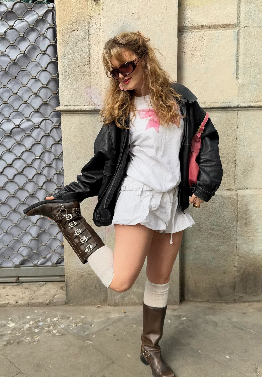 Giubbotto di pelle nera, t-shirt bianca con design rosa, pantaloni corti bianchi, stivali marroni al ginocchio con fibbie e calzini a ginocchio color crema.