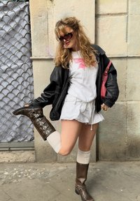 Giubbotto di pelle nera, t-shirt bianca con design rosa, pantaloni corti bianchi, stivali marroni al ginocchio con fibbie e calzini a ginocchio color crema.