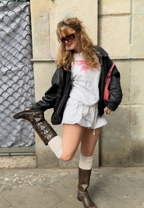 Chaqueta de cuero negra, camiseta blanca con diseño rosa, pantalones cortos blancos, botas marrones hasta la rodilla con hebillas y medias crema hasta la rodilla.