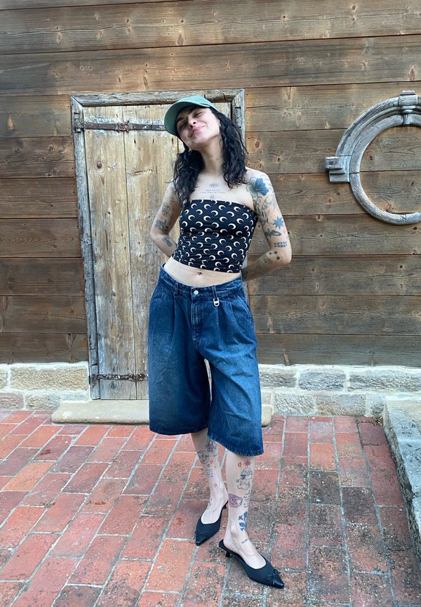 Jeune femme tatouée portant un top tube à motif lune, un short en jean large, une casquette verte et des chaussures pointues noires, debout sur un pavé en briques.