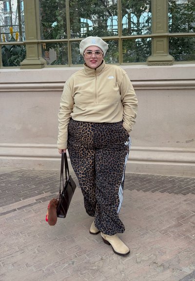 Persona con forro polar marrón claro, pantalones con estampado de leopardo, botas beige, boina beige y gafas, sosteniendo un bolso marrón, de pie en un camino pavimentado.