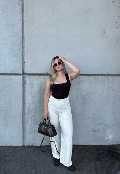 Mujer con camiseta negra, pantalones blancos, zapatos negros y gafas de sol, sosteniendo un bolso negro, de pie frente a una pared de hormigón gris.