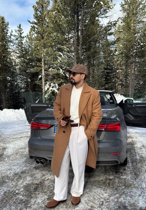 Hombre con abrigo marrón, pantalones blancos y gorra, de pie sosteniendo guantes junto a un coche gris con las puertas abiertas en un entorno forestal nevado.