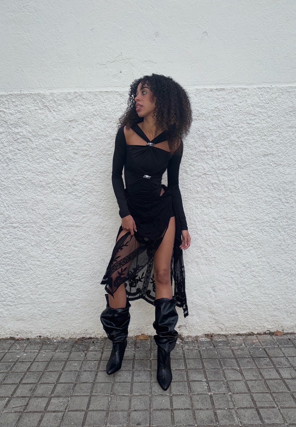 Svart langermet topp med rynkedetaljer og sølvaksenter, lagdelt med et gjennomsiktig svart skjørt med blomsterbroderi og høy splitt. Sorte knelange støvler.