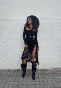 Svart langermet topp med rynkedetaljer og sølvaksenter, lagdelt med et gjennomsiktig svart skjørt med blomsterbroderi og høy splitt. Sorte knelange støvler.