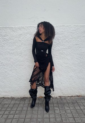 Top negro de manga larga con diseño fruncido y detalles plateados, combinado con una falda negra transparente con bordado floral y una abertura alta. Botas negras hasta la rodilla.