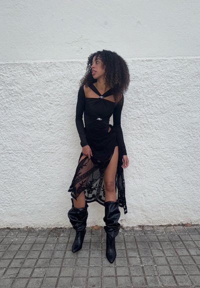 Top negro de manga larga con diseño fruncido y detalles plateados, combinado con una falda negra transparente con bordado floral y una abertura alta. Botas negras hasta la rodilla.