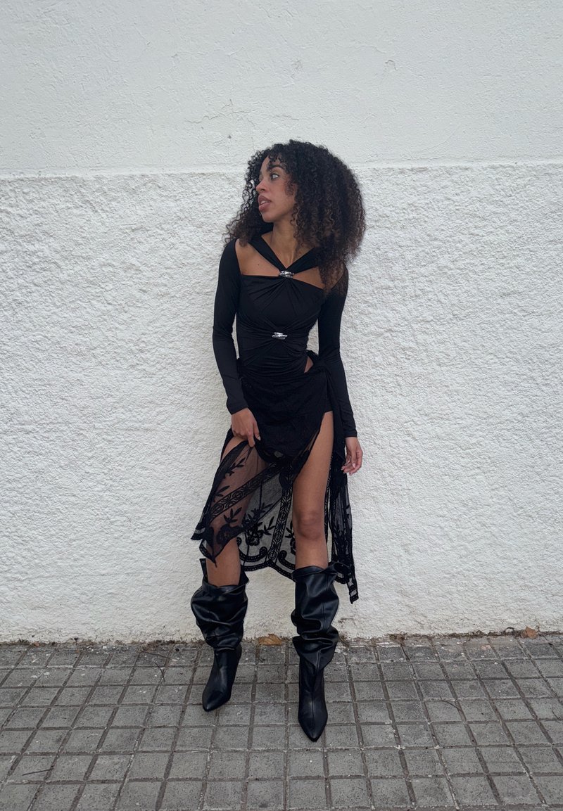 Juodas ilgomis rankovėmis palaidinė su rauktu dizainu ir sidabriniais akcentais, derinama su permatoma juoda sijonu, puoštame gėlių išsiuvinėjimu ir aukšta skeltimi. Juodi ilgi batai iki kelių.