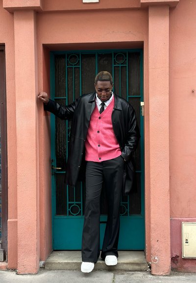 Abrigo de cuero negro, cárdigan rosa con botones, pantalones de vestir negros y zapatos blancos de suela gruesa. De pie junto a una puerta teal con una pared rosa.