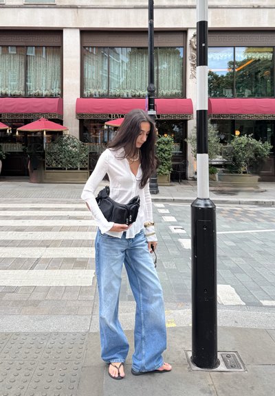 Camisa blanca de manga larga con cuello abotonado, jeans de pierna ancha azul claro, bolso negro bandolera y chanclas. Poste de calle a rayas cerca.