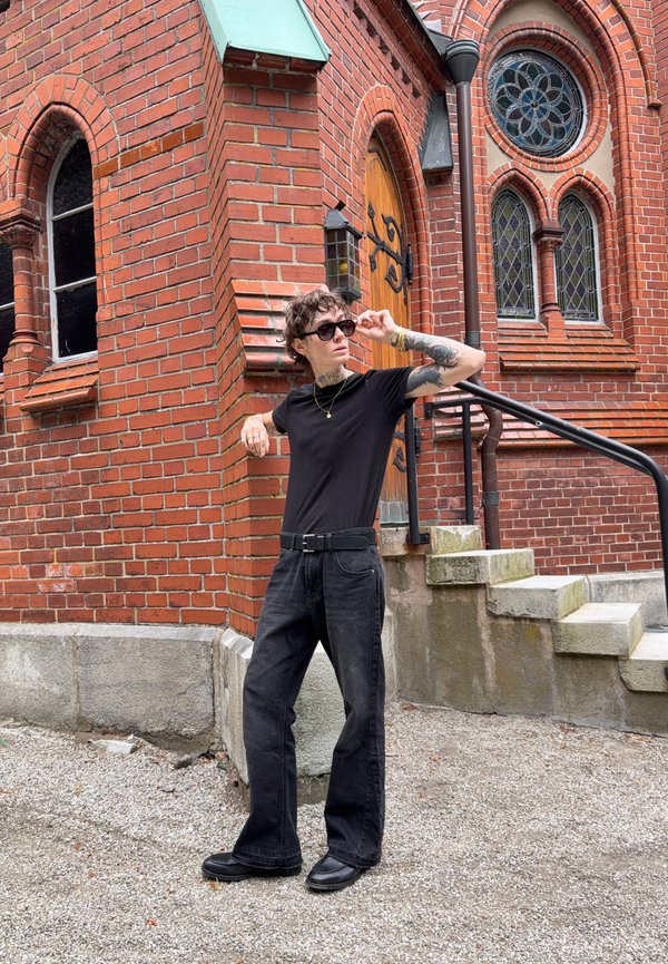 Person with curly hair and tattoos wears black clothes and sunglasses, standing by red brick building with stained glass windows and stone steps.