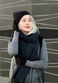 Fekete kötött sapka, szürke sál és szürke bordás pulóver csipkés részletekkel, fekete bőr mellény fölött, egy modern háttér előtt.