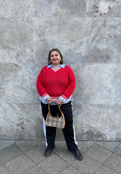 Suéter rojo de punto con rayas blancas, superpuesto a una camisa azul, combinado con pantalones negros que tienen rayas blancas y una pequeña bolsa de cuadros marrón.