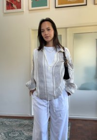 Cămașă din in cu dungi albastre și albe, design cu decolteu în V, asociată cu pantaloni albi largi. Accesoriile includ o geantă de umăr neagră.