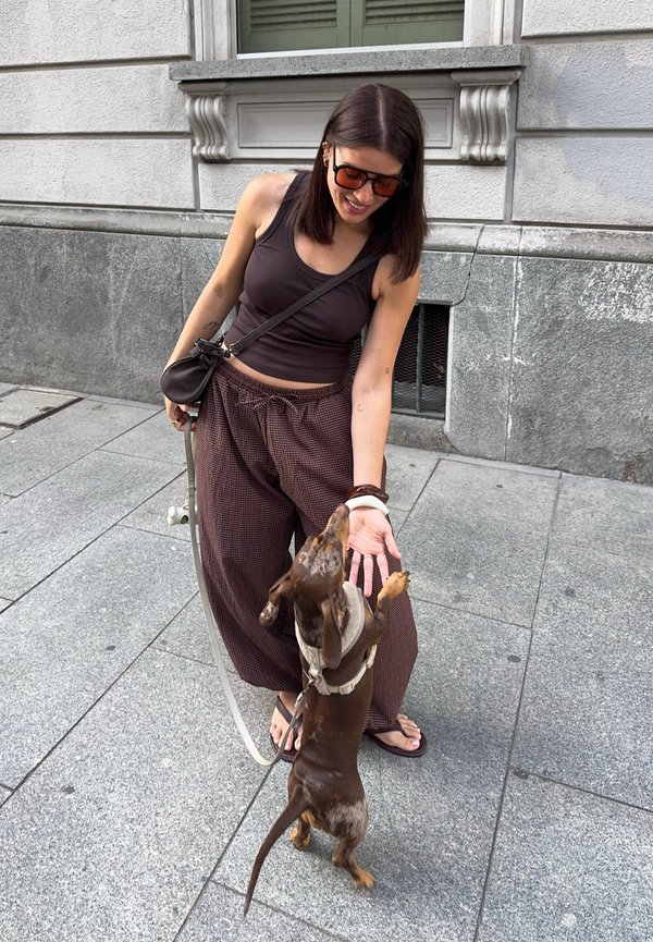 Mujer con gafas de sol y pantalones holgados sonriendo mientras extiende la mano hacia un pequeño dachshund marrón y beige que está de pie sobre sus patas traseras en la acera.