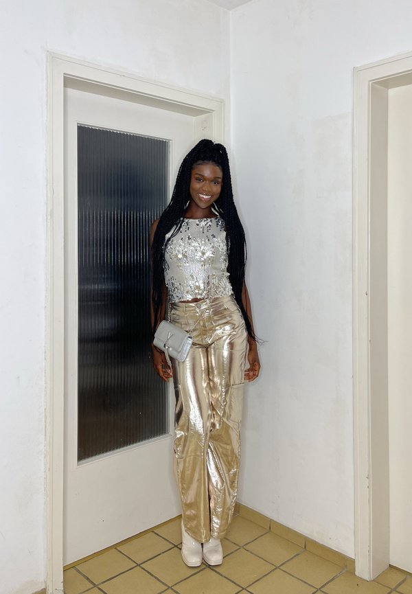 Svetlikav srebrni crop top s sequinci, metalne zlate hlače širokega kroja in bele petke; drži majhno srebrno torbico.