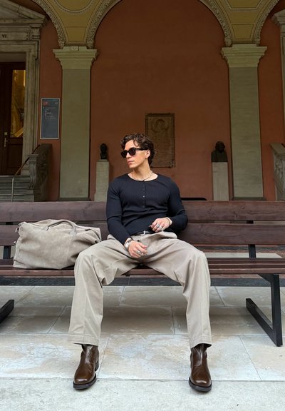 Camisa negra de manga larga con rayas, pantalones de pierna ancha color beige, botas de cuero marrón, joyería de plata y un bolso de lona gris sobre un banco de madera.