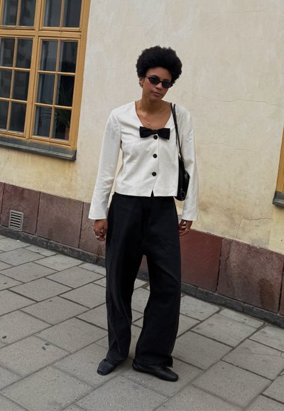 Chaqueta de lino blanca con un lazo negro y botones, combinada con pantalones negros holgados. Los accesorios incluyen gafas de sol oscuras y un bolso negro.