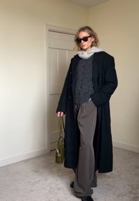 Lungo cappotto nero, maglione grigio e pantaloni color sabbia. Accessori includono una borsa verde e occhiali da sole neri. Tessuti lisci e colori neutri.