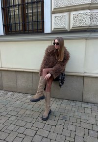 Femme portant un grand manteau en fourrure marron, des lunettes de soleil, une jupe à carreaux et des bottes beige montant jusqu'aux genoux, assise sur un mur bas dans une rue pavée.