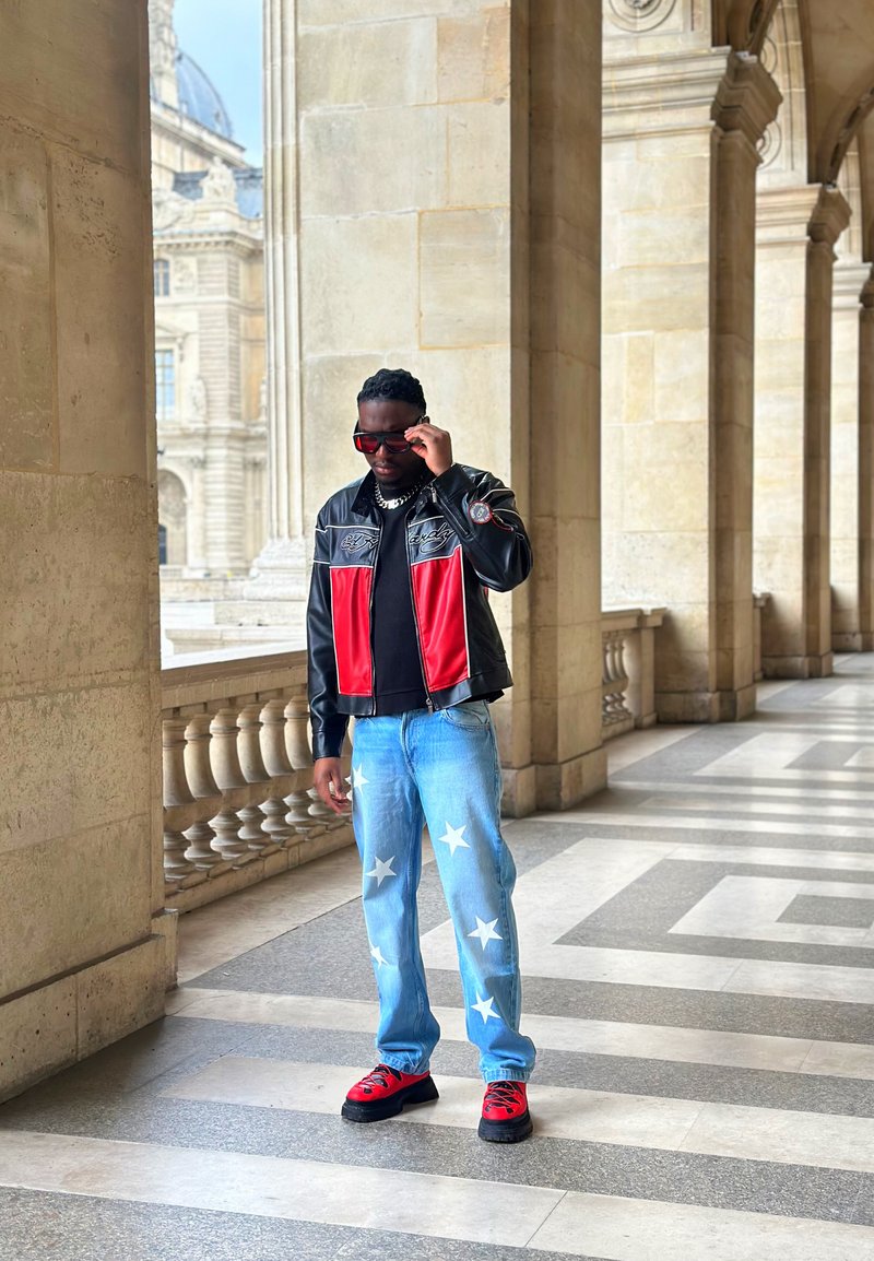 Homme ajustant ses lunettes de soleil, portant une veste noire et rouge, un jean bleu à motif étoiles et des chaussures rouges, debout dans un passage bordé de colonnes en pierre.
