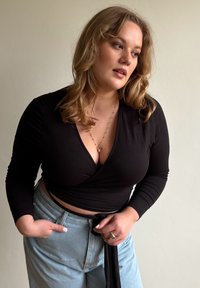 Juodas ilgomis rankovėmis suveržiamas viršus su gilia V formos iškirpte, derinamas su šviesiai mėlynais aukšto liemens džinsais. Modelis viena ranka laiko kišenę, plaukai paleisti laisvai.