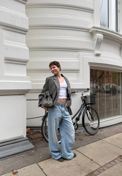 Mujer con blazer gris, camiseta blanca sin mangas y jeans anchos apoyada en una bicicleta negra junto a un edificio blanco curvo con grandes ventanas.