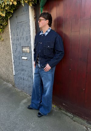 Joven con gafas de sol, chaqueta de mezclilla oscura, camisa de cuadros, pantalones anchos azules y botas negras, de pie frente a una puerta de madera roja al aire libre.