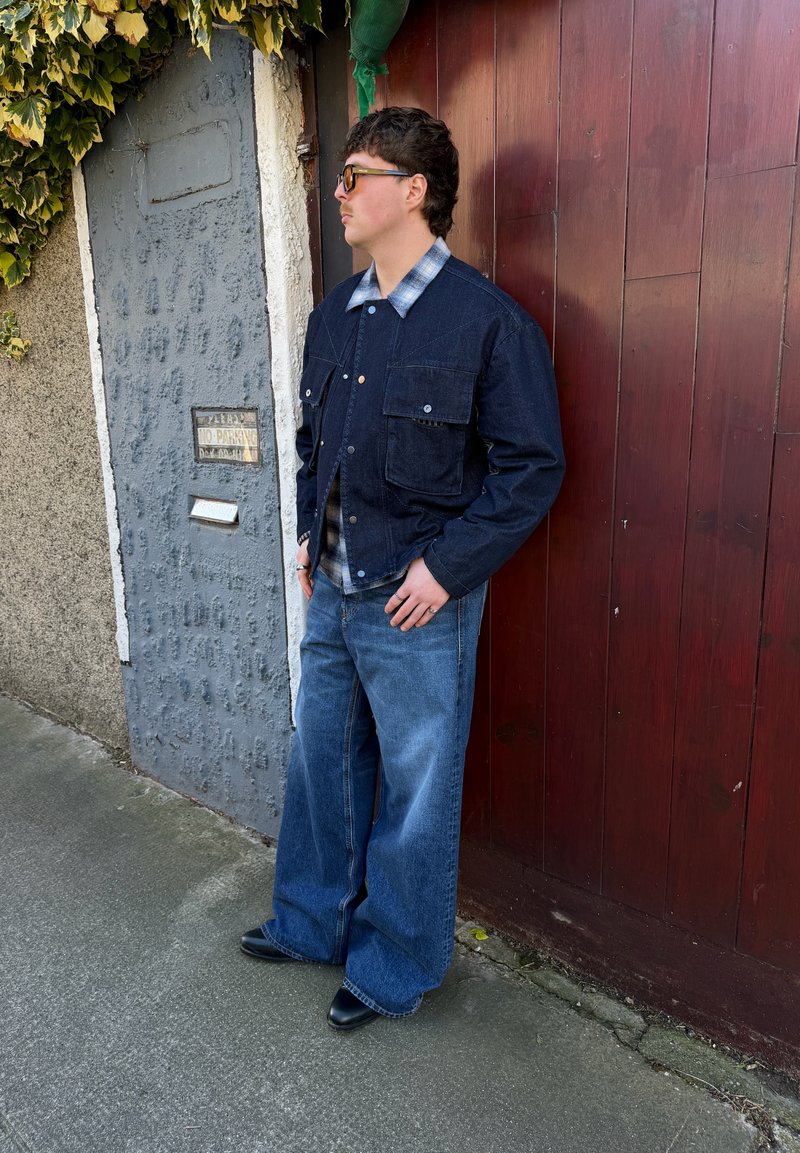 Joven con gafas de sol, chaqueta de mezclilla oscura, camisa de cuadros, pantalones anchos azules y botas negras, de pie frente a una puerta de madera roja al aire libre.