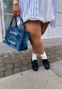 Τσάντ�α από τζιν με λευκό λογότυπο "SAM SøE", συνδυασμένη με μαύρες λοφάδες και φραμπαλά λευκές κάλτσες, κάτω από ριγέ πουκαμίσα. Υπόβαθρο από πέτρες.