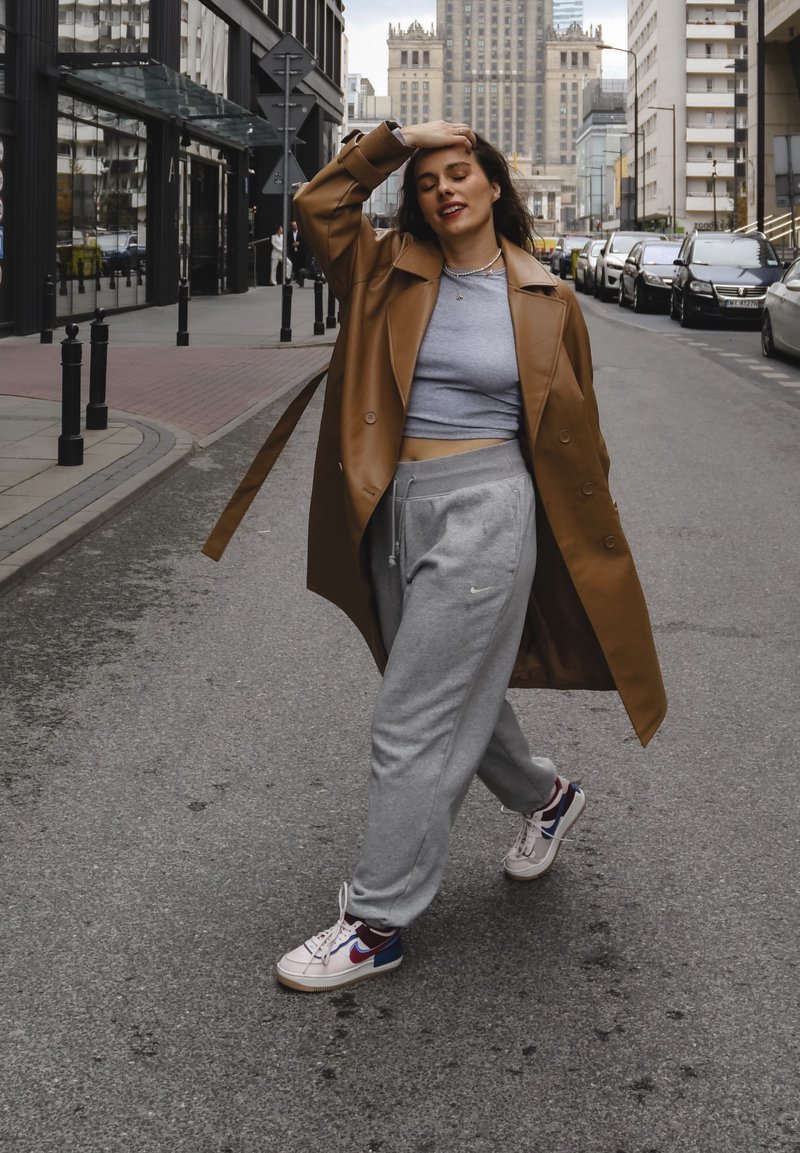 Καφετί παλτό πάνω από γκρι cropped top και φόρμες. Λευκά αθλητικά παπούτσια με κόκκινες και μπλε λεπτομέρειες. Αστική σκηνή με παρκαρισμένα αυτοκίνητα.