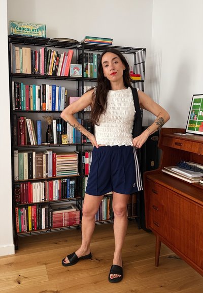 Top blanco texturizado, pantalones cortos azul marino con rayas blancas, sandalias negras de tipo slide. Muebles de madera y estanterías en el fondo.