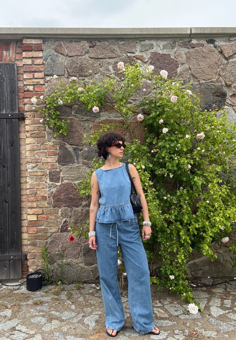 Personne portant un haut sans manches en denim bleu et un pantalon, des lunettes de soleil noires et des sandales, debout près d'un mur en pierre avec une rose grimpante.