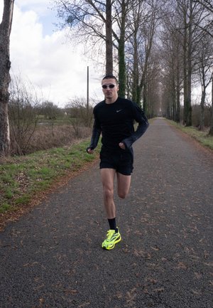 Hombre corriendo por un sendero bordeado de árboles, vestido con ropa deportiva negra y zapatillas de correr de color amarillo neón, bajo un cielo nublado.