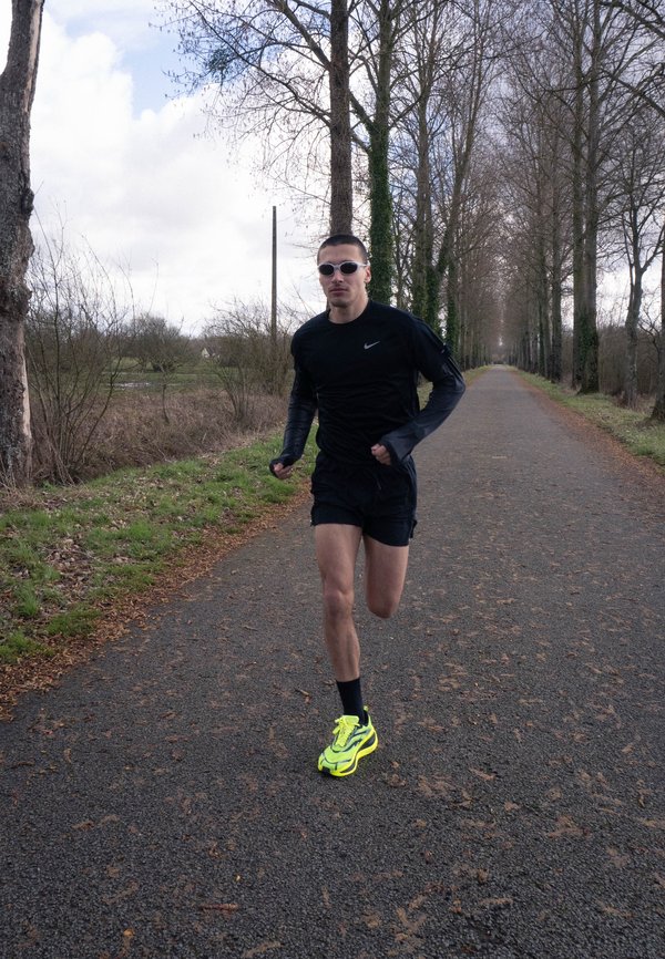 Mann läuft auf einem von Bäumen gesäumten Weg, trägt schwarze Sportkleidung und neongelbe Laufschuhe unter einem bewölkten Himmel.