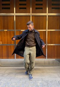 Un homme ajuste son manteau noir ouvert, portant un pull en laine marron à torsades, un pantalon olive et des baskets bleues, debout contre un mur en bois lambrissé.